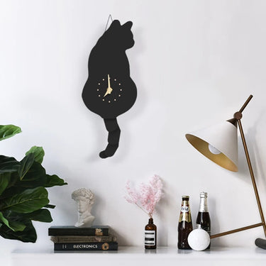 The Playful Purring Wall Cat Clock by KittyNook Cat Co. hangs above a desk with books, plants, bottles, and a lamp.
